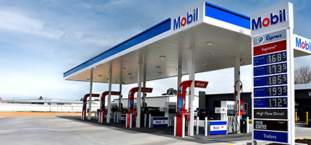 Mobil-branded fuel station with multiple fuel pumps and a digital price board displaying fuel prices. The canopy is blue and white with the Mobil logo, and the station has a clean, organized layout under an overcast sky.