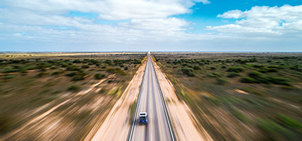 Aerial view of a single car driving along a long, straight highway cutting through a flat, open landscape with sparse vegetation. The scene conveys a sense of solitude and vast distance, under a partly cloudy sky..