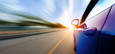 Close-up view of a blue car driving on an open road. The camera captures the vehicle from a low angle beside the side mirror, with motion blur indicating speed. The background shows a bright, sunlit sky with a blurred horizon.